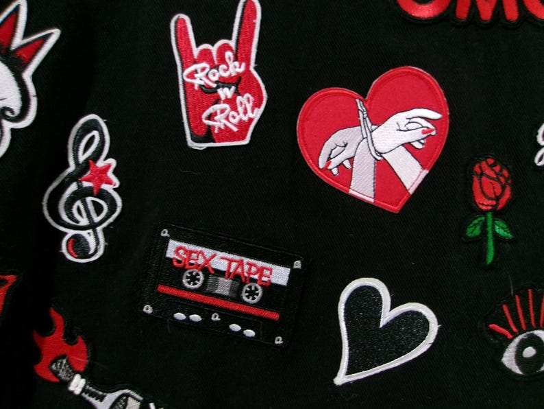May include: A black fabric surface decorated with various embroidered patches including a red hand gesture with the text Rock n Roll, a red heart with bound hands, a cassette tape labeled SEX TAPE, a treble clef, a red rose, and a black heart.