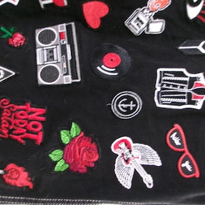 May include: A black fabric garment adorned with various embroidered patches, including a boombox, a red rose, a vinyl record, a guitar with wings, red sunglasses, a heart, and text that reads Not Today Satan in bold red lettering.
