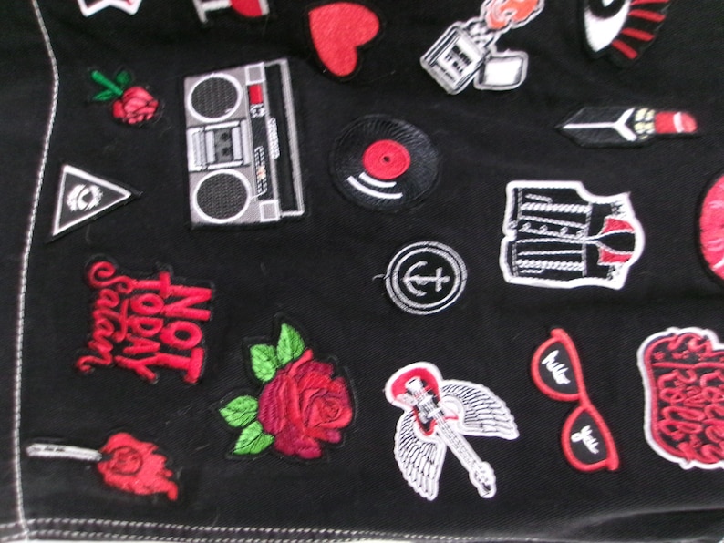 May include: A black fabric garment adorned with various embroidered patches, including a boombox, a red rose, a vinyl record, a guitar with wings, red sunglasses, a heart, and text that reads Not Today Satan in bold red lettering.