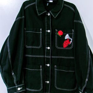 May include: A black denim jacket featuring prominent white contrast stitching, a classic collar, and a button-down front. The jacket includes four front pockets, with the upper left pocket decorated with an embroidered red rose and a red anatomical heart.
