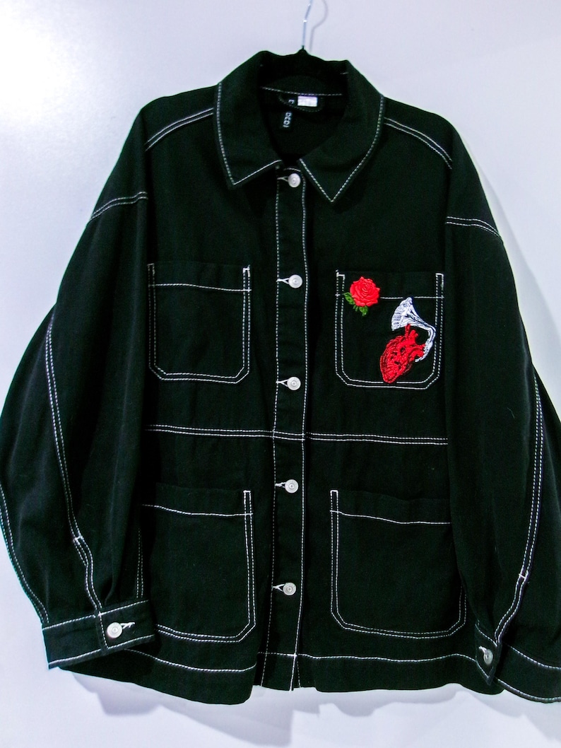 May include: A black denim jacket featuring prominent white contrast stitching, a classic collar, and a button-down front. The jacket includes four front pockets, with the upper left pocket decorated with an embroidered red rose and a red anatomical heart.