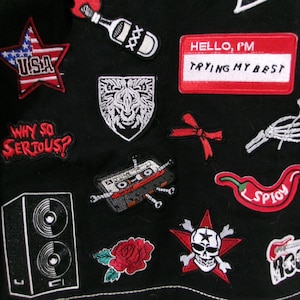 May include: A black fabric garment adorned with various embroidered patches, including a red star with USA text, a white lion shield, a red name tag reading HELLO, I'M TRYING MY BEST, a cassette tape, a red chili pepper, and a skull with crossbones.