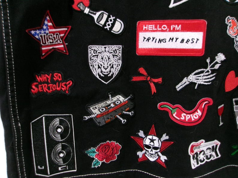 May include: A black fabric garment adorned with various embroidered patches, including a red star with USA text, a white lion shield, a red name tag reading HELLO, I'M TRYING MY BEST, a cassette tape, a red chili pepper, and a skull with crossbones.