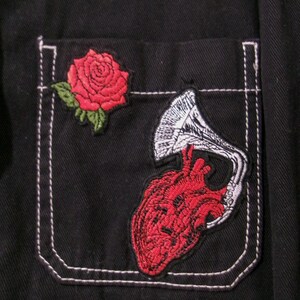 May include: A black shirt pocket featuring two embroidered patches. One patch is a red rose with green leaves. The second patch depicts a red anatomical heart connected to a white and black gramophone horn, stitched onto the dark fabric.