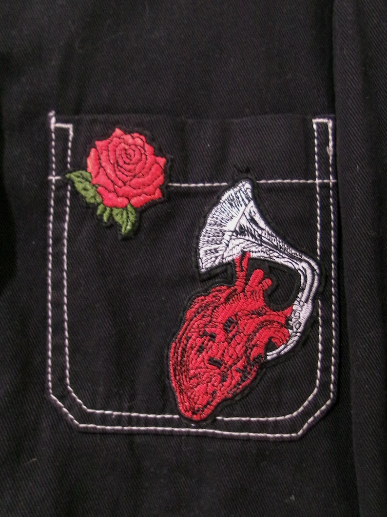 May include: A black shirt pocket featuring two embroidered patches. One patch is a red rose with green leaves. The second patch depicts a red anatomical heart connected to a white and black gramophone horn, stitched onto the dark fabric.