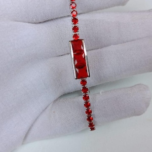 May include: A silver bracelet featuring a series of small, round red gemstones and a central rectangular section with larger red stones. The jewelry is displayed on a white surface.