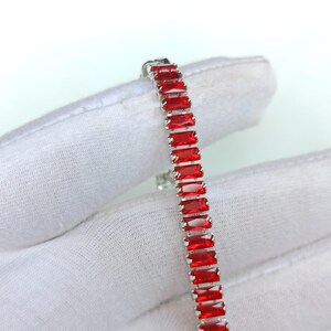 Può includere: Un bracciale d'argento con pietre preziose rettangolari di colore rosso vivo. Le pietre sono incastonate vicine, creando una linea continua di colore. Il bracciale è esposto su uno sfondo bianco, evidenziando il design e il colore del gioiello.