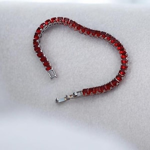 May include: A silver bracelet with a row of square, red gemstones. The bracelet is unclasped, showing the clasp mechanism. The gemstones are a vibrant red color, and the bracelet is set against a white background.