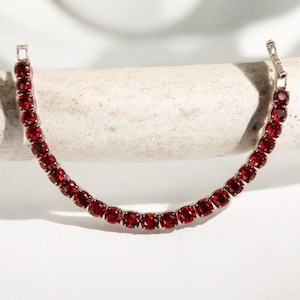 May include: A silver tennis bracelet with a continuous line of vivid, round, red gemstones. The bracelet is presented on a neutral surface against a white backdrop, emphasizing the jewellery's colour and design.