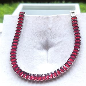 May include: A silver bracelet featuring a row of vibrant, rectangular red gemstones. The gemstones are set closely together, creating a continuous line of color. The bracelet is displayed on a white surface.
