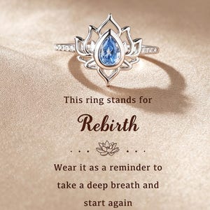 Puede incluir: Un anillo de tono plateado con una piedra azul en forma de pera en el centro, engastada en un marco de metal inspirado en un loto. El texto dice: This ring stands for Rebirth. Wear it as a reminder to take a deep breath and start again.