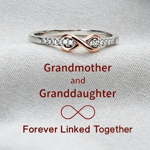May include: A silver-toned ring featuring a central rose gold infinity symbol accented with small clear stones. The text below reads: Grandmother and Granddaughter, an infinity symbol, and the phrase Forever Linked Together.