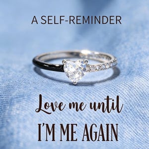 Puede incluir: Un anillo de tono plateado con una piedra preciosa transparente en forma de corazón en el centro, con un lado de la banda en negro pulido y el otro engastado con pequeñas piedras transparentes. Se muestra el texto A SELF-REMINDER Love me until I'M ME AGAIN.
