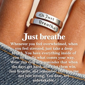 May include: A silver-toned wrap ring featuring two flat bands. The top band is engraved with a small lotus flower icon and the word "Just," while the bottom band is engraved with the word "Breathe" in a black script font.