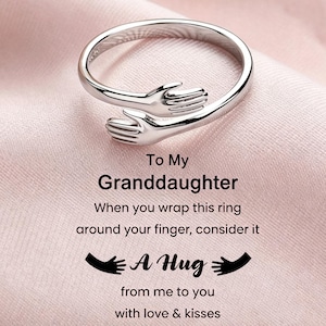 Puede incluir: Un anillo de metal plateado con dos manos que se encuentran en el centro para formar un abrazo. El texto dice: To My Granddaughter, When you wrap this ring around your finger, consider it A Hug from me to you with love & kisses.