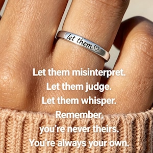 Può includere: Un anello in metallo color argento con il testo inciso "let them" seguito da un piccolo simbolo a forma di cuore. L'anello è indossato su un dito e l'immagine include il testo sovrapposto: "Let them misinterpret. Let them judge. Let them whisper. Remember, you're never theirs. You're always your own."