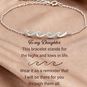 May include: A silver chain bracelet featuring a central wave-shaped charm accented with small light blue stones. The text reads: To my Daughter, This bracelet stands for the highs and lows in life. Wear it as a reminder that I will be there for you through them all.