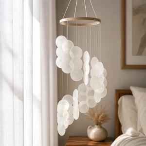 Op de afbeelding: Een witte windgong van capiz-schelpen met een houten ring aan de bovenkant. De windgong heeft talrijke ronde, doorschijnende schelpen die aan touwtjes zijn opgehangen. Het ontwerp is eenvoudig en elegant, geschikt voor binnendecoratie.
