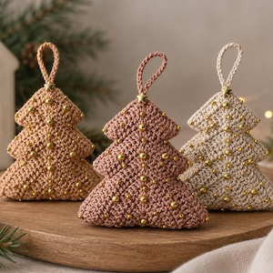 May include: Three triangular crochet Christmas tree ornaments in tan, dusty rose, and cream colors. Each tree features a small gold star at the top, a matching crochet hanging loop, and scattered gold beads across the textured yarn surface.