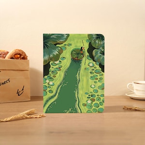 Pode incluir: Um caderno retangular com uma ilustração vibrante de uma pessoa em pé sobre uma grande folha de vitória-régia num lago verde, usando uma vara longa para navegar através de água cheia de inúmeras folhas de lírio e pequenas flores brancas.