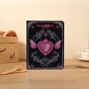 May include: Black notebook with pink heart and wing design. The cover features the text "DEATH NOTE" in pink, with decorative swirls and a heart-shaped emblem. The notebook is on a wooden surface.