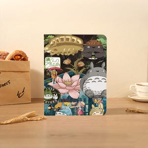 May include: A colorful notebook featuring various Totoro characters and imagery. The design includes a large gray Totoro, a pink lotus flower, and other characters from the animated film. The background is a mix of green and blue.