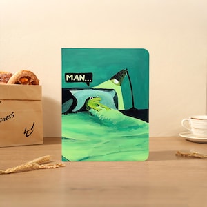 Pode incluir: Um cartão de felicitações retangular com uma ilustração pintada de um sapo verde deitado na cama sob um cobertor verde. Um balão de fala acima do sapo contém o texto "MAN..." e uma luminária de mesa verde ilumina a cena em tons de verde.