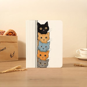 May include: A white notebook featuring a stack of five cartoon cat faces in black, orange, blue, and gray. The cats peek over the edge of the notebook. The notebook is on a wooden surface with a paper bag and a cup in the background.
