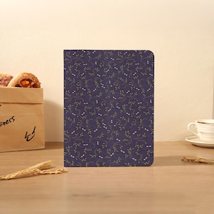 May include: A rectangular notebook with a dark purple cover featuring a repeating pattern of thin green vines and small white dragonfly silhouettes. The notebook has rounded corners and is positioned upright on a flat surface.