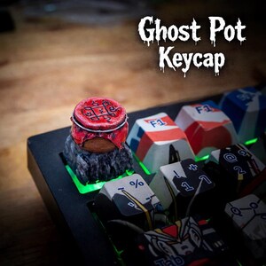 May include: A custom keycap, designed as a ghost pot, is red and black and sits on a mechanical keyboard. Other keys are visible. The text "Ghost Pot Keycap" is above the keycap. The keyboard has green lighting.