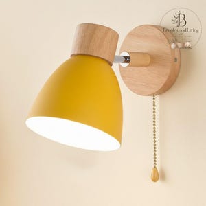 May include: A yellow wall-mounted lamp with a wooden base and accents. The lamp features a rounded, cone-shaped shade and a pull chain. The wooden base is circular and the lamp is mounted on a light-colored wall.