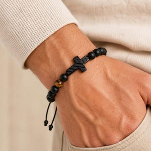 Puede incluir: Pulsera de cuentas negras con un colgante de cruz negra. La pulsera incluye una cuenta de ojo de tigre y un cierre de cordón negro ajustable. La pulsera se lleva en la muñeca.