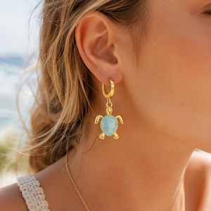 Puede incluir: Un pendiente de aro dorado con un dije colgante de tortuga marina. La tortuga tiene un marco de oro pulido y una incrustación de piedra veteada de color azul claro en su caparazón. El pendiente cuenta con un cierre de bisagra seguro.