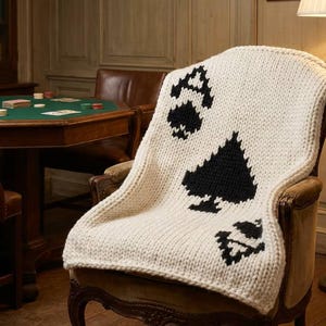 May include: A chunky knit throw blanket in cream white featuring a large black spade suit symbol from a deck of cards. The blanket is draped over a wooden armchair and displays a prominent black spade icon centered on the thick, textured yarn fabric.
