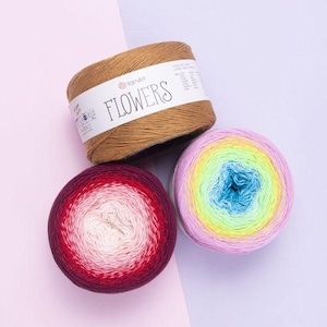 Può includere: Tre gomitoli di filato YarnArt Flowers. Un gomitolo color marrone chiaro presenta un'etichetta bianca con la scritta FLOWERS. Un altro gomitolo mostra una sfumatura dal bordeaux scuro al bianco. Il terzo gomitolo ha un vivace gradiente dal viola al giallo neon.