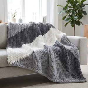 Könnte beinhalten: Eine grob gestrickte Decke in Grau- und Weißtönen liegt über einem grauen Sofa. Die Decke hat ein geometrisches Muster mit diagonalen Streifen. Das dicke Garn verleiht der Decke ein gemütliches, strukturiertes Aussehen, perfekt, um Wärme und Stil zu verleihen.