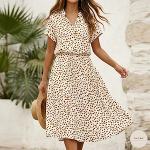 Pode incluir: Vestido creme com padrão de manchas castanhas. O vestido apresenta mangas curtas, gola e cintura com cordão. Um chapéu de palha é segurado na mão esquerda. O vestido é até o joelho e tem uma saia fluida.