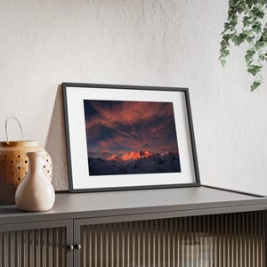 Könnte beinhalten: Ein gerahmtes Landschaftsfoto mit schneebedeckten Berggipfeln, die von einem leuchtend rosa und orangefarbenen Sonnenuntergangshimmel mit dunklen Wolken beleuchtet werden, in einem dünnen schwarzen Rahmen mit weißem Passepartout auf einem dunkelgrauen Schrank.