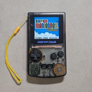 Puede incluir: Una consola Game Boy Color transparente que muestra "Super Mario Bros. Deluxe". El dispositivo portátil tiene una correa de muñeca amarilla. La pantalla muestra una escena de juego colorida. El texto "©1985, 1999 Nintendo" y "Game Boy Color" son visibles.