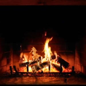Puede incluir: Una chimenea acogedora con un fuego crepitante. Las llamas son de color naranja brillante y amarillo, iluminando los troncos y el marco de piedra. El hogar es oscuro, creando un ambiente cálido y acogedor.