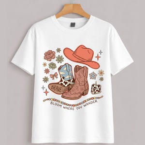 May include: A white short-sleeved t-shirt featuring a graphic print of a coral cowboy hat, two cowboy boots, flowers, a small cake, and stars. One boot has a cow print and the word BRAVE. The text reads BLOOM WHERE YOU WANDER below the items.