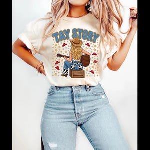 Pu&ograve; includere: Una t-shirt color crema con la grafica di una persona dai lunghi capelli biondi che indossa un cappello da cowboy e suona la chitarra seduta su una cassa di legno. Il testo TAY STORY &egrave; stampato sopra, circondato da piccoli cappelli rossi.