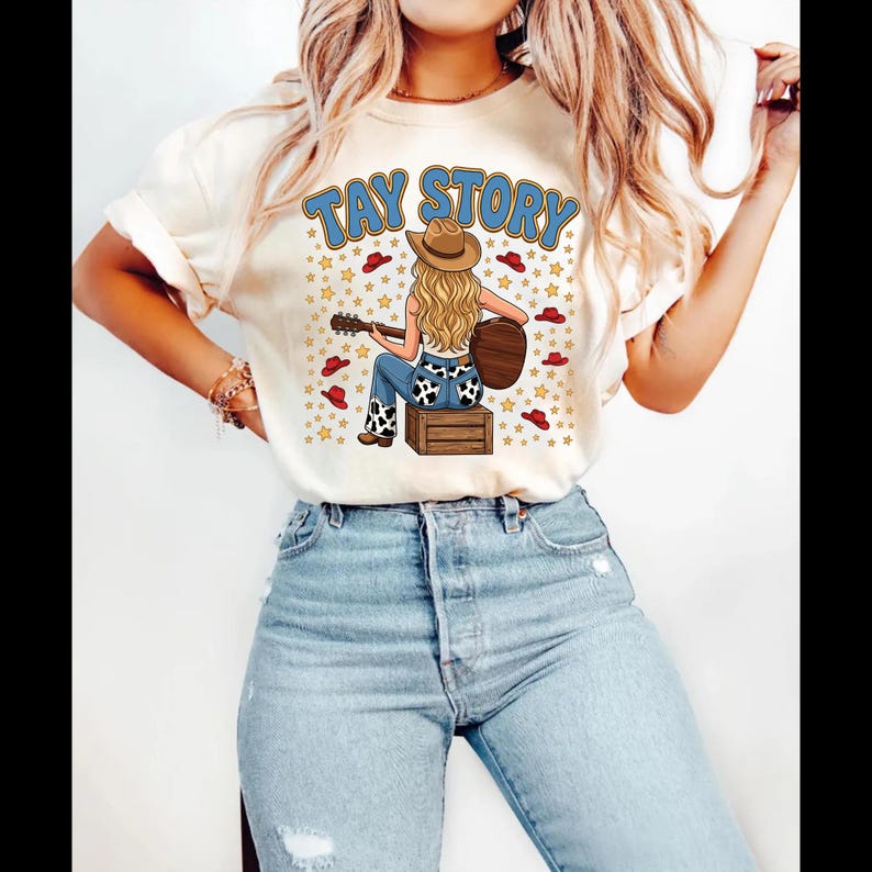 Pu&ograve; includere: Una t-shirt color crema con la grafica di una persona dai lunghi capelli biondi che indossa un cappello da cowboy e suona la chitarra seduta su una cassa di legno. Il testo TAY STORY &egrave; stampato sopra, circondato da piccoli cappelli rossi.