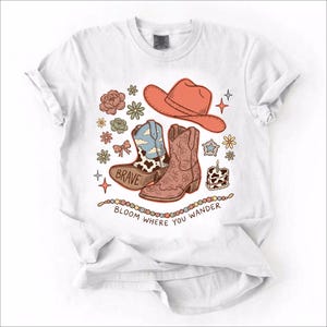 May include: A white t-shirt featuring a graphic print of a coral cowboy hat, two cowboy boots, flowers, a star, and a small cake. One boot has a cow print and the word BRAVE. Below the graphics, a string of beads sits above the text BLOOM WHERE YOU WANDER.