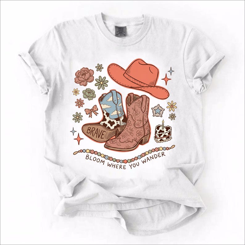 May include: A white t-shirt featuring a graphic print of a coral cowboy hat, two cowboy boots, flowers, a star, and a small cake. One boot has a cow print and the word BRAVE. Below the graphics, a string of beads sits above the text BLOOM WHERE YOU WANDER.