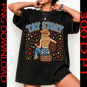 Pu&ograve; includere: Una t-shirt nera con lavaggio minerale che presenta la grafica di una persona bionda con cappello da cowboy seduta su una cassa di legno mentre suona la chitarra. Il testo TAY STORY appare in blu sopra la figura, circondato da stelle gialle e cappelli rossi.
