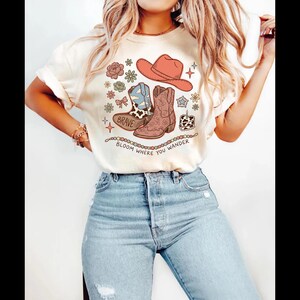 May include: A cream-coloured short-sleeve t-shirt featuring a graphic print of a cowboy hat, two cowboy boots, flowers, and a small cake. The text "BRAVE" is on one boot, and the text "BLOOM WHERE YOU WANDER" is printed in an arc below the design.