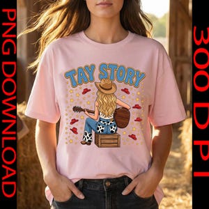 Pu&ograve; includere: Una t-shirt rosa chiaro con la grafica di una persona bionda che indossa un cappello da cowboy e jeans con stampa mucca, seduta su una cassa di legno mentre suona una chitarra acustica. Il testo TAY STORY &egrave; stampato sopra, circondato da stelle gialle e cappelli rossi.