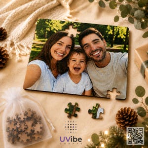 May include: A rectangular jigsaw puzzle featuring a colour photograph of a smiling woman, a young child, and a man outdoors. The puzzle is partially assembled, with two loose pieces placed below the main image and a small white drawstring bag containing additional puzzle pieces to the left.
