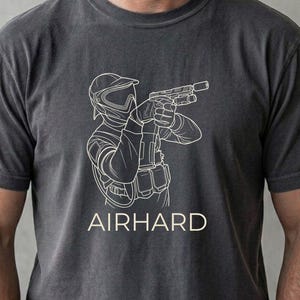 May include: Dark gray t-shirt with a white outline graphic of a person in tactical gear holding a gun. The word "AIRHARD" is printed in white below the graphic. The shirt is made of a soft material.